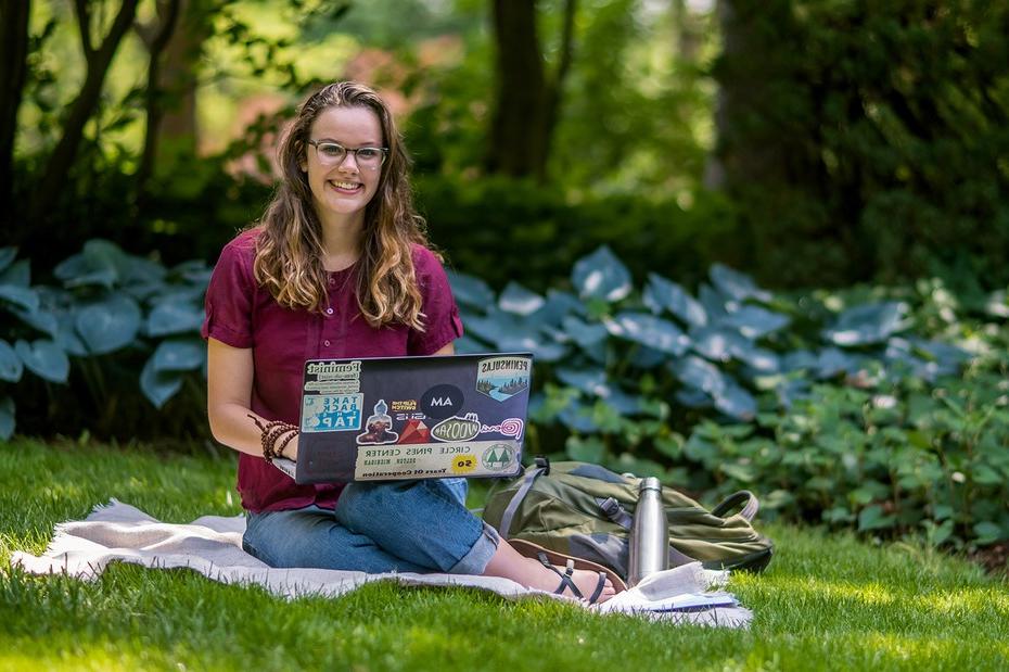 学生坐在草坪上的毯子上，拿着笔记本电脑微笑